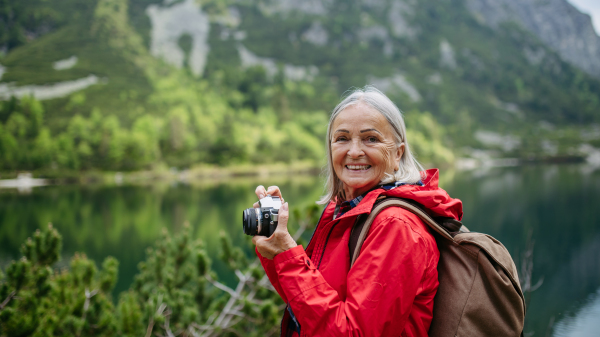 Potrait of active senior woman hiking in autumn mountains, on senior friendly trail. Senior tourist with backpack taking pictures with analog camera.