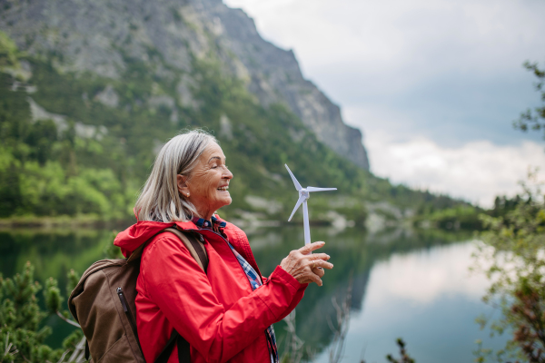 Potrait of active female senior tourist holding a model of wind turbine. Senior woman with backpack spinning the blade of turbine. Concept of renewable, green energy and sustainable lifestyle.