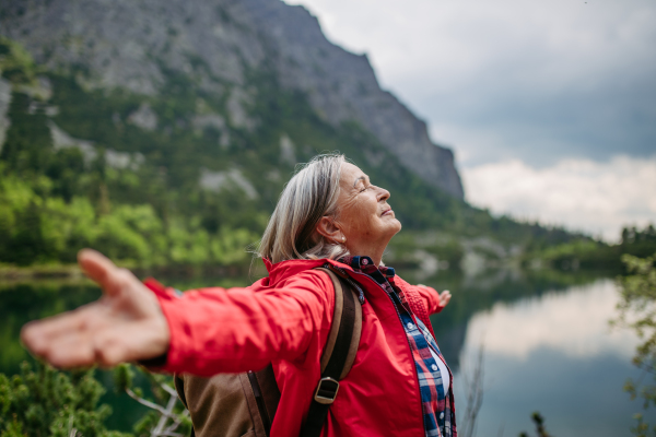 Waist up shot of happy elderly woman with hands in the air. Senior tourist feeling satisfied after hike in autumn mountains.