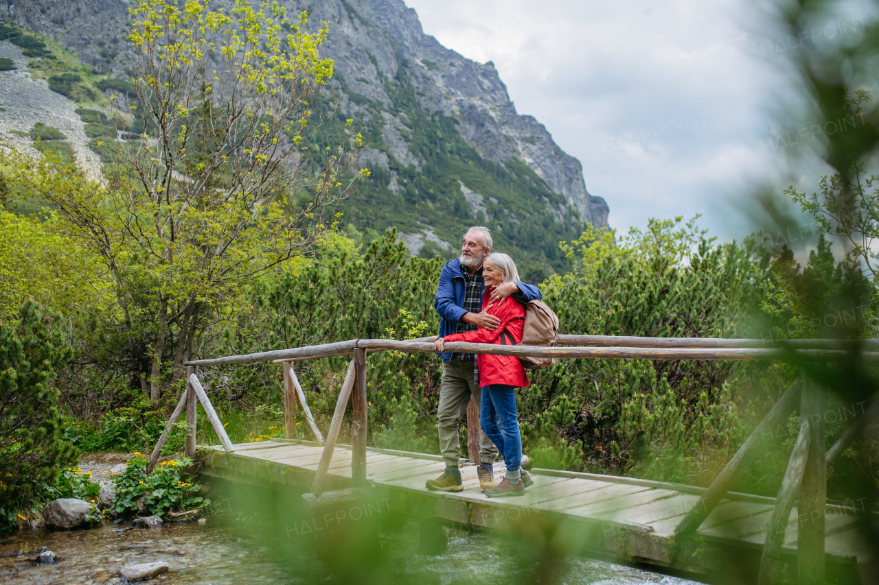 Portrait of beautiful active elderly couple hiking together in autumn mountains, standing on wooden bridge. Senior spouses on the vacation in the mountains celebrating anniversary. Senior tourists with backpacks using trekking poles for stability.