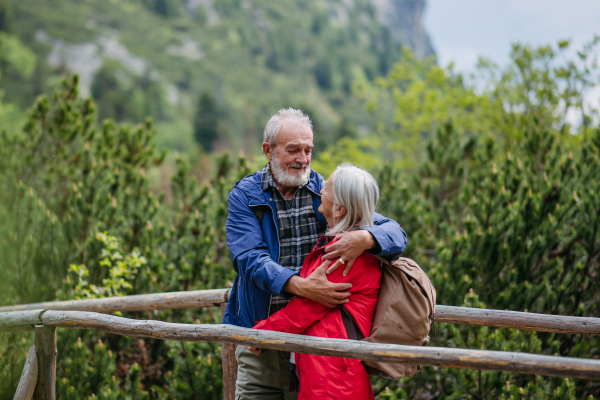 Portrait of beautiful active elderly couple hiking together in autumn mountains, standing on wooden bridge. Senior spouses on the vacation in the mountains celebrating anniversary. Senior tourists are embracing on a wooden bridge during a hike.