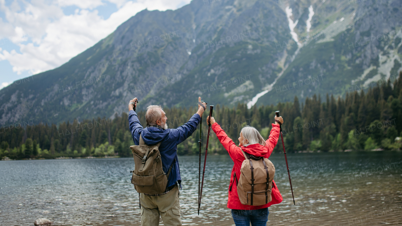 Rear view of active elderly couple hiking together in autumn mountains, on senior friendly trail. Senior spouses on the vacation in the mountains celebrating anniversary. Senior tourists with backpacks using trekking poles for stability.