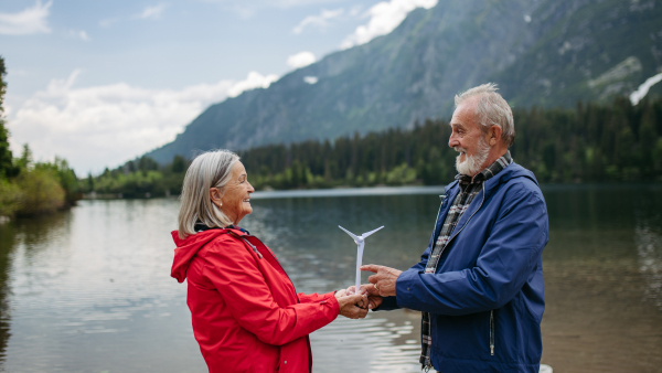 Portrait of senior couple holding a model of wind turbine, standing in the middle stunning nature. Concept of renewable, green energy and sustainable lifestyle.