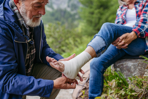 Senior man bandaging the injured leg of the female tourist. A senior woman injured her arm during hike in the mountains. Tourist went off-trail and fell.