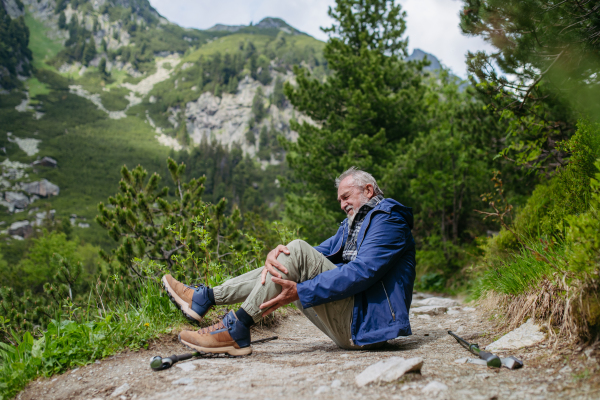 Senior man injured his leg during hike in the mountains. Tourist went off-trail and fell. Elderly tourist resting on the ground, feeling pain under his kneecap, also known as Hiker's knee.