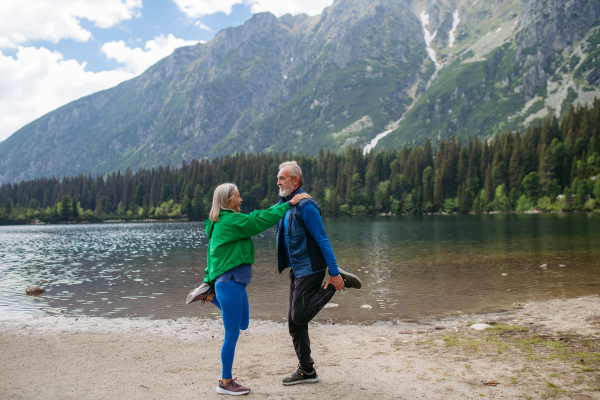 Full body shot of senior couple doing outdoor yoga, tai chi, pilates by the lake in the autumn. Elderly husband and wife spending active vacation in the mountains, enjoying combination of physical activity and relaxation. Pensioners stretching after outdoor workout