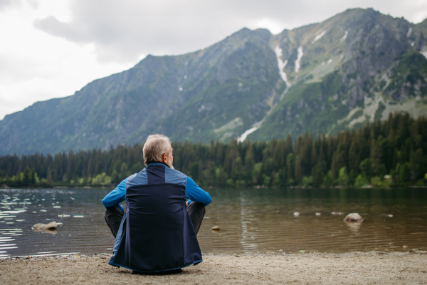 Rear view of active senior man resting after hiking in autumn mountains. Thoughtful elderly man sitting on the shore, enjoying view of untouched nature.