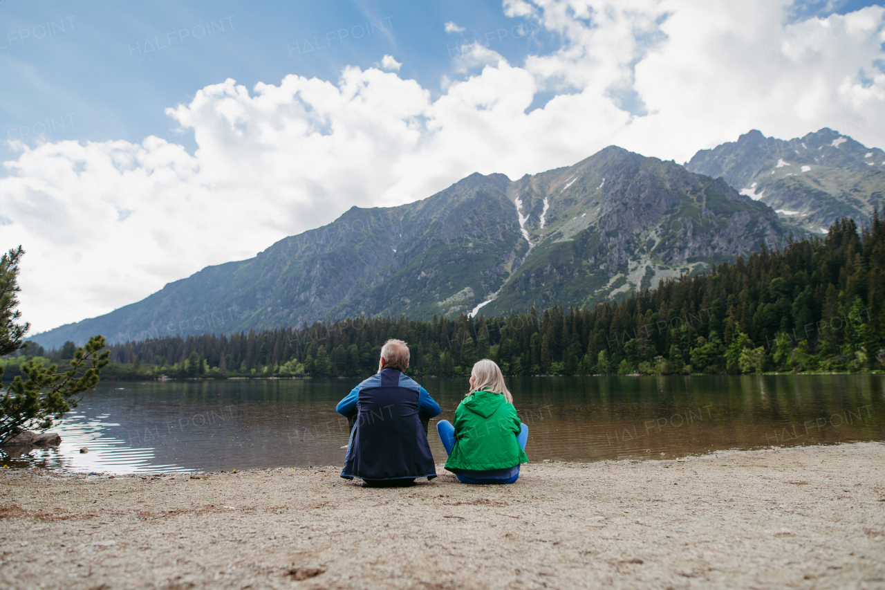 Rear view of active senior couple resting after hiking in autumn mountains. Thoughtful elderly man and woman sitting on the shore, enjoying view of untouched nature.