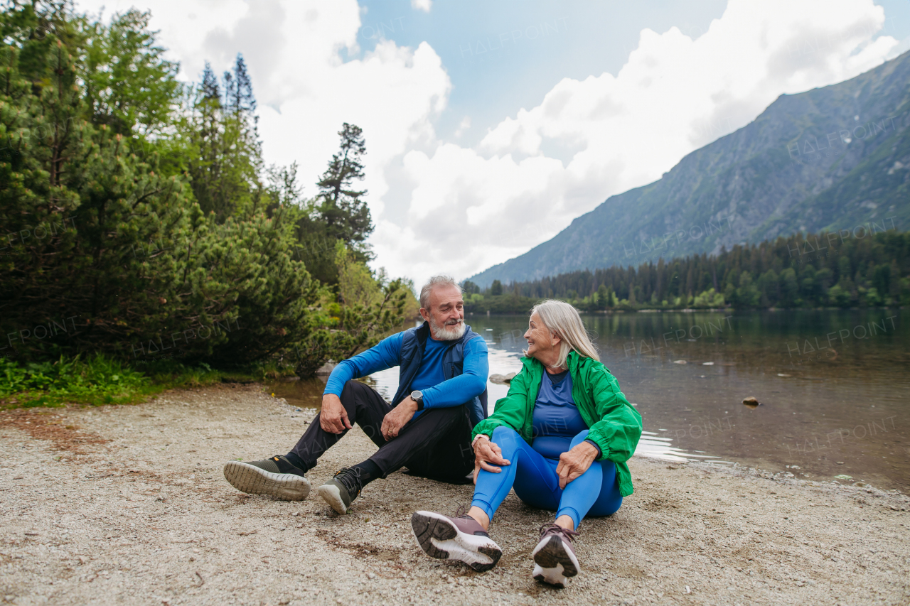 Active senior couple resting and looking at each other lovingly after hiking in autumn mountains. Thoughtful elderly man and woman sitting on the shore, enjoying view of untouched nature.