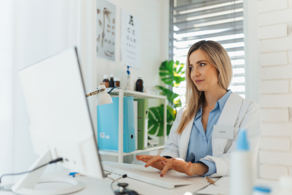 Young beautiful female doctor working on laptop in doctor's office. Physician doing paperwork and administrative tasks.