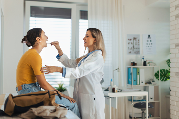 Doctor examining young patient's sore throat, looking into the throat using a tongue depressor.