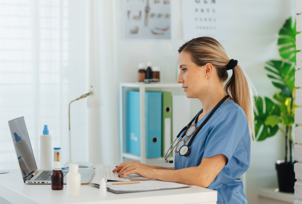 Side view of beautiful female doctor working on laptop in doctor's office. Physician doing paperwork and administrative tasks.