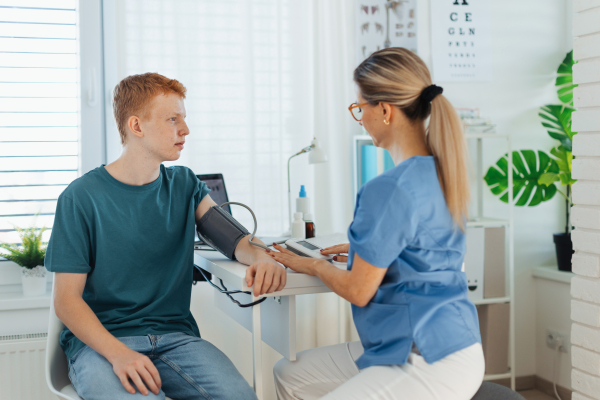 Doctor, nurse measuring boy's blood pressure, using blood pressure monitor. Teenage boy visiting paediatrician for annual preventive physical examination. Concept of preventive health care for adolescents.