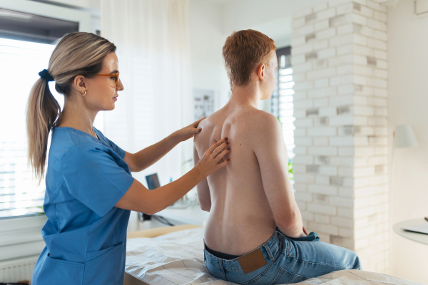 An orthopedist examines the spine, posture, and spinal deformities of a teenage patient. Teenage boy visiting paediatrician for annual preventive physical examination. Concept of preventive health care for adolescents.