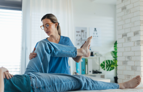 Physiotherapist doing manual therapy, exercise with teenage boy's knee, rehabilitation. Orthopedist examining and treating painful injured knee of young athlete. Concept of teen preventive health care.