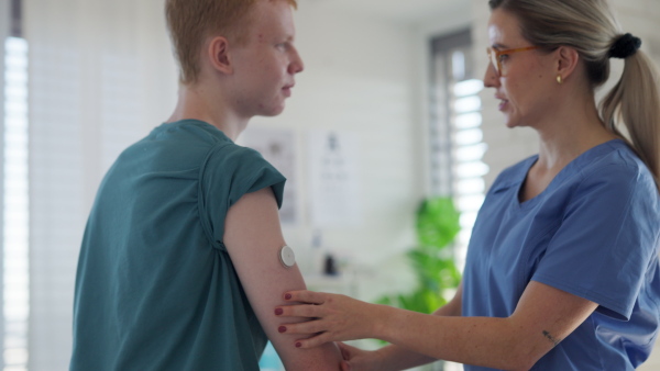 Diabetologist doctor, nurse applying a continuous glucose monitor sensor on arm of a diabetic teenager. CGM device making life of teenage boy easier, helping manage his illness and focus on other activities.