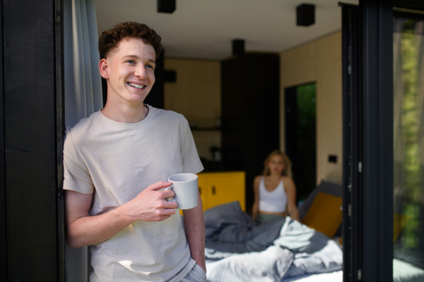 Young man stading in terrace with cup of coffee and looking away, his girlfrend sitting in a bed.