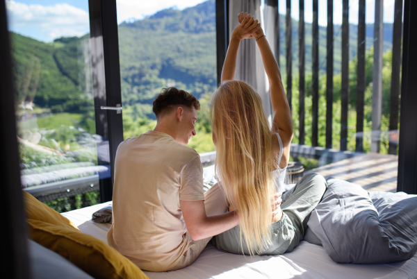 A rear view of young couple in love sitting on bed bed in morning in their new home in tiny house, sustainable living concept.