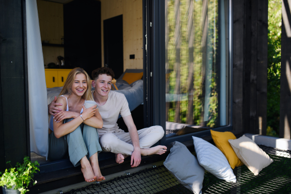 A young couple sitting and cuddling with cup of coffee in terrace in their new home in tiny house in woods, sustainable living concept.