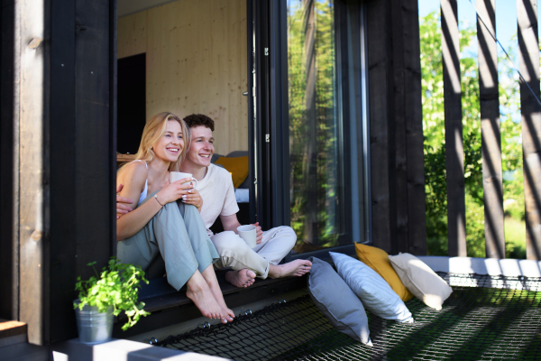 A young couple sitting and cuddling with cup of coffee in terrace in their new home in tiny house in woods, sustainable living concept.