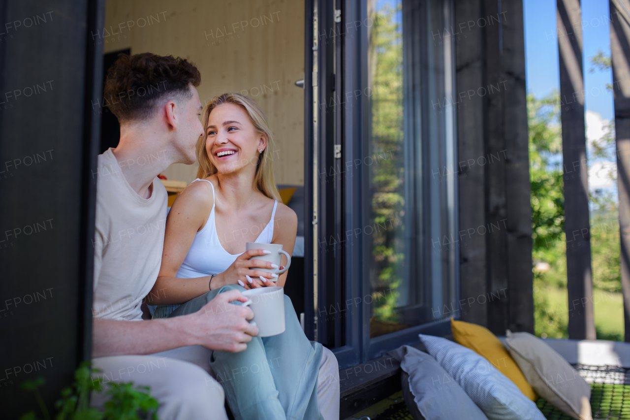 A young couple sitting and cuddling in hammock terrace in their new home in tiny house in woods, sustainable living concept.