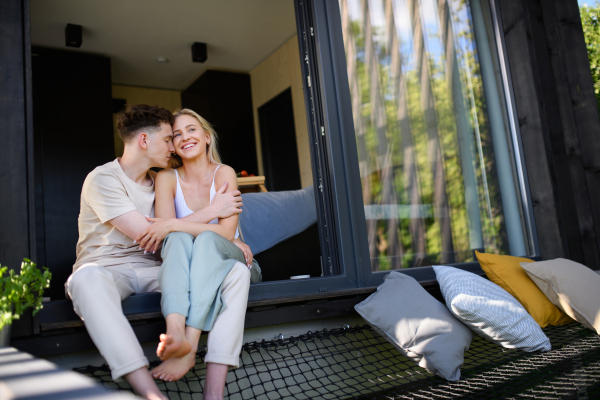 A young couple sitting and cuddling in hammock terrace in their new home in tiny house in woods, sustainable living concept.