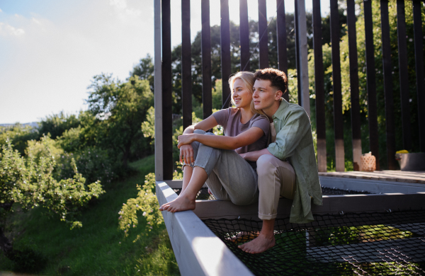 A young couple sitting and cuddling in hammock terrace in their new home in tiny house in woods, sustainable living concept.