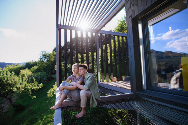 A young couple sitting and cuddling in hammock terrace in their new home in tiny house in woods, sustainable living concept.