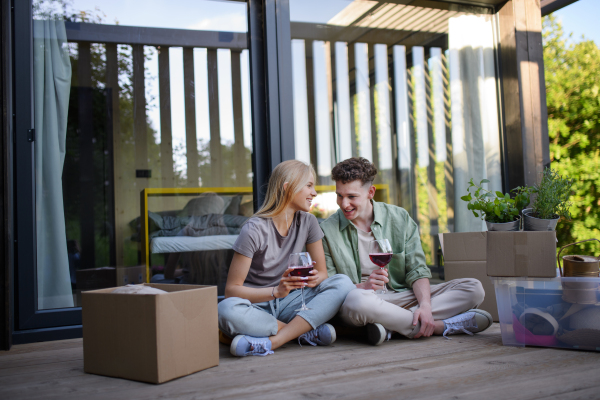 A cheerful young couple moving in their new tiny house in woods sitting on floor on terrace and drinking wine. Conception of moving and sustainable living.