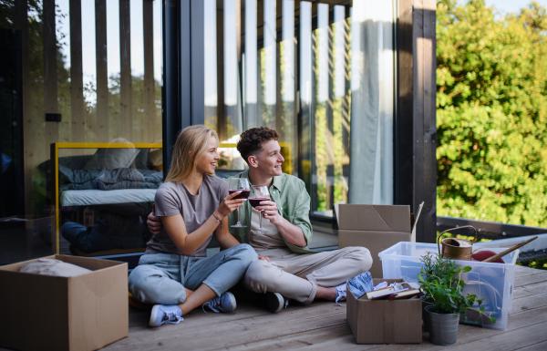 A cheerful young couple moving in their new tiny house in woods sitting on floor on terrace and drinking wine. Conception of moving and sustainable living.