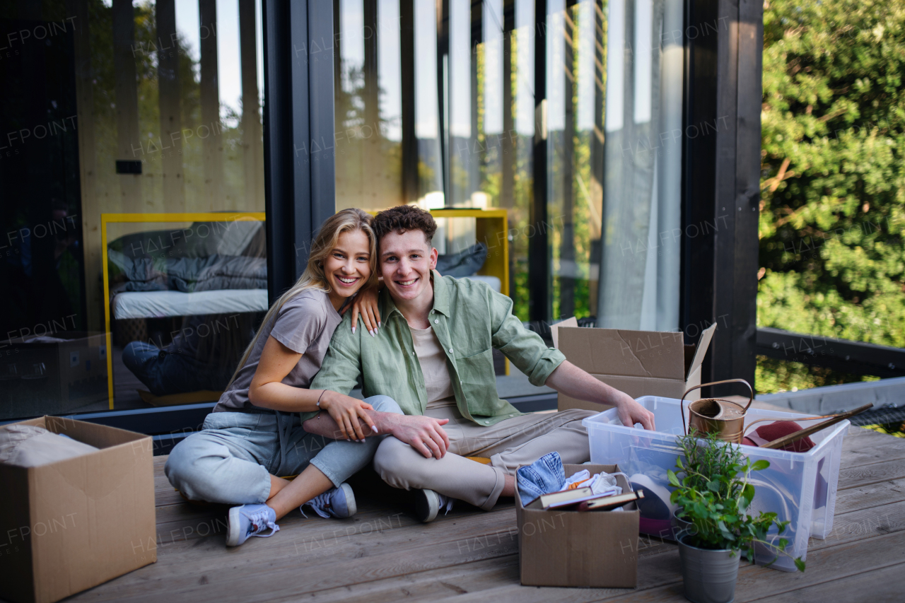 A cheerful young couple moving in their new tiny house in woods sitting on floor on terrace. Conception of moving and sustainable living.