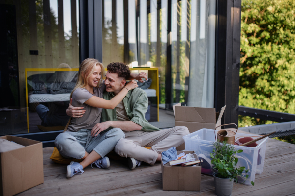 A cheerful young couple moving in their new tiny house in woods sitting on floor on terrace. Conception of moving and sustainable living.