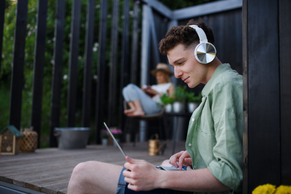 A happy young couple with laptop and headphones resting outdoors in a tiny house, weekend away and remote office concept.