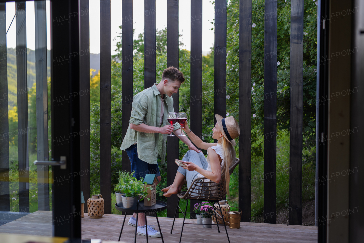 A young couple clinking with wine outdoors, weekend away in tiny house in countryside, sustainable living.