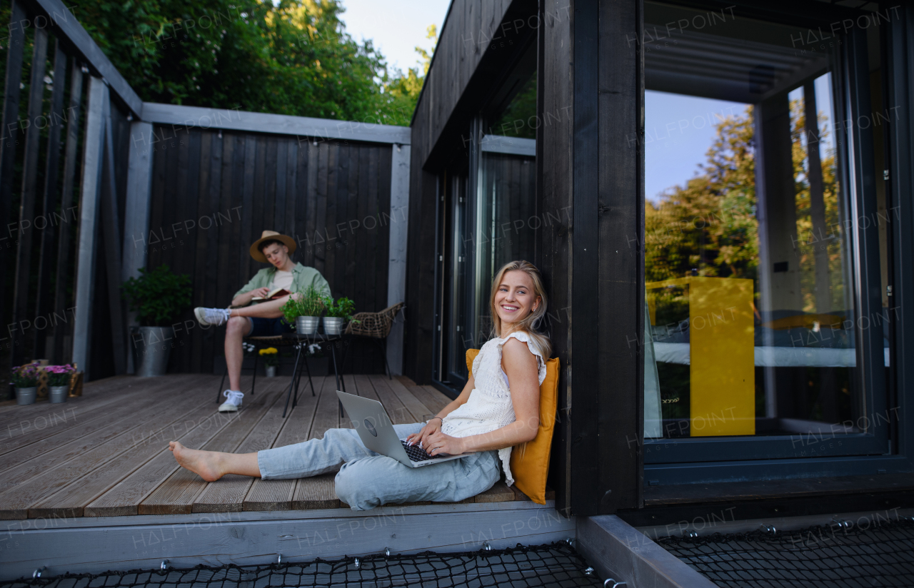 A happy young couple with laptop resting outdoors in a tiny house, weekend away and remote office concept.