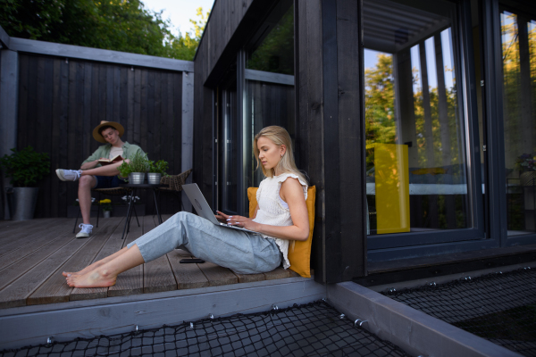 A happy young couple with laptop resting outdoors in a tiny house, weekend away and remote office concept.