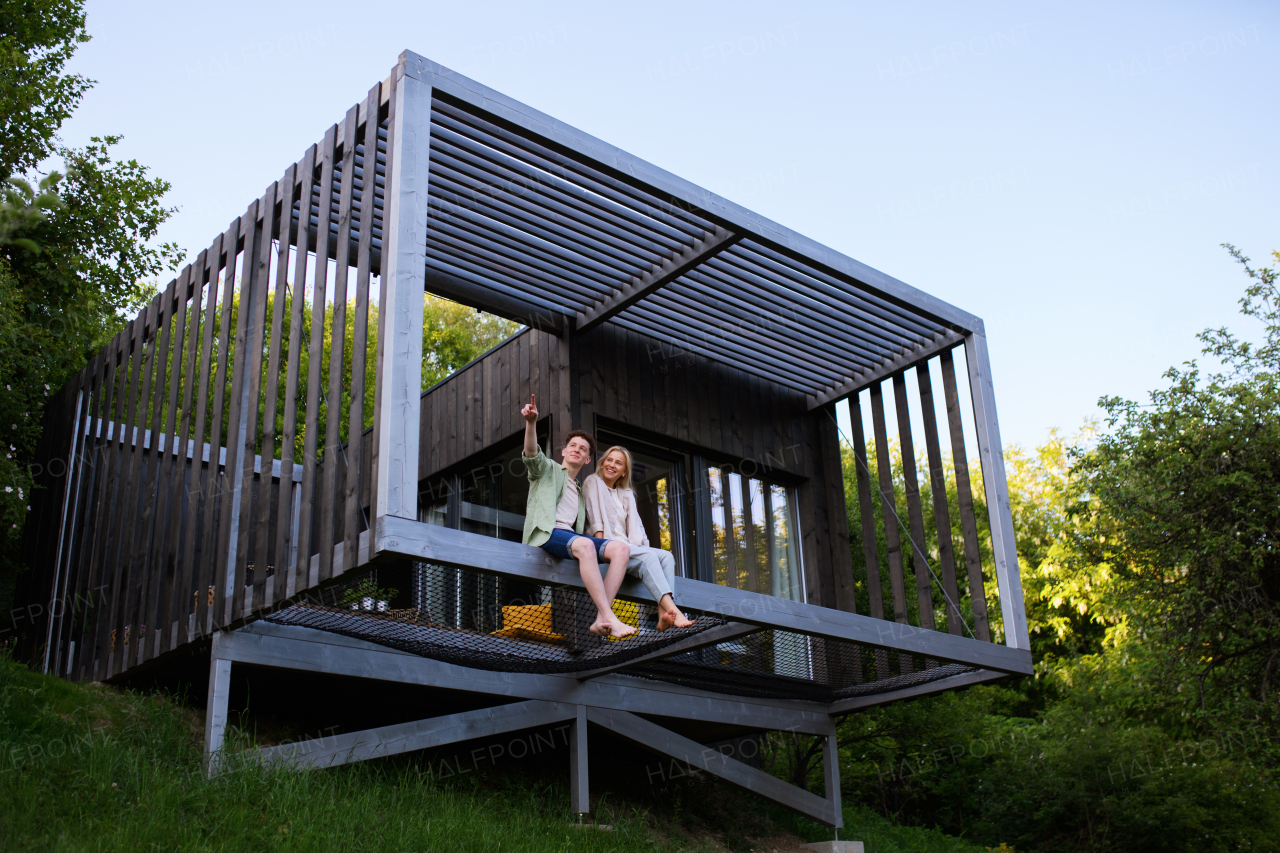 A young couple sitting and cuddling in hammock terrace in their new home in tiny house in woods, sustainable living concept.