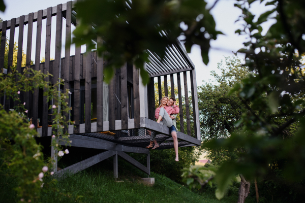 A young couple sitting and cuddling in hammock terrace in their new home in tiny house in woods, sustainable living concept.