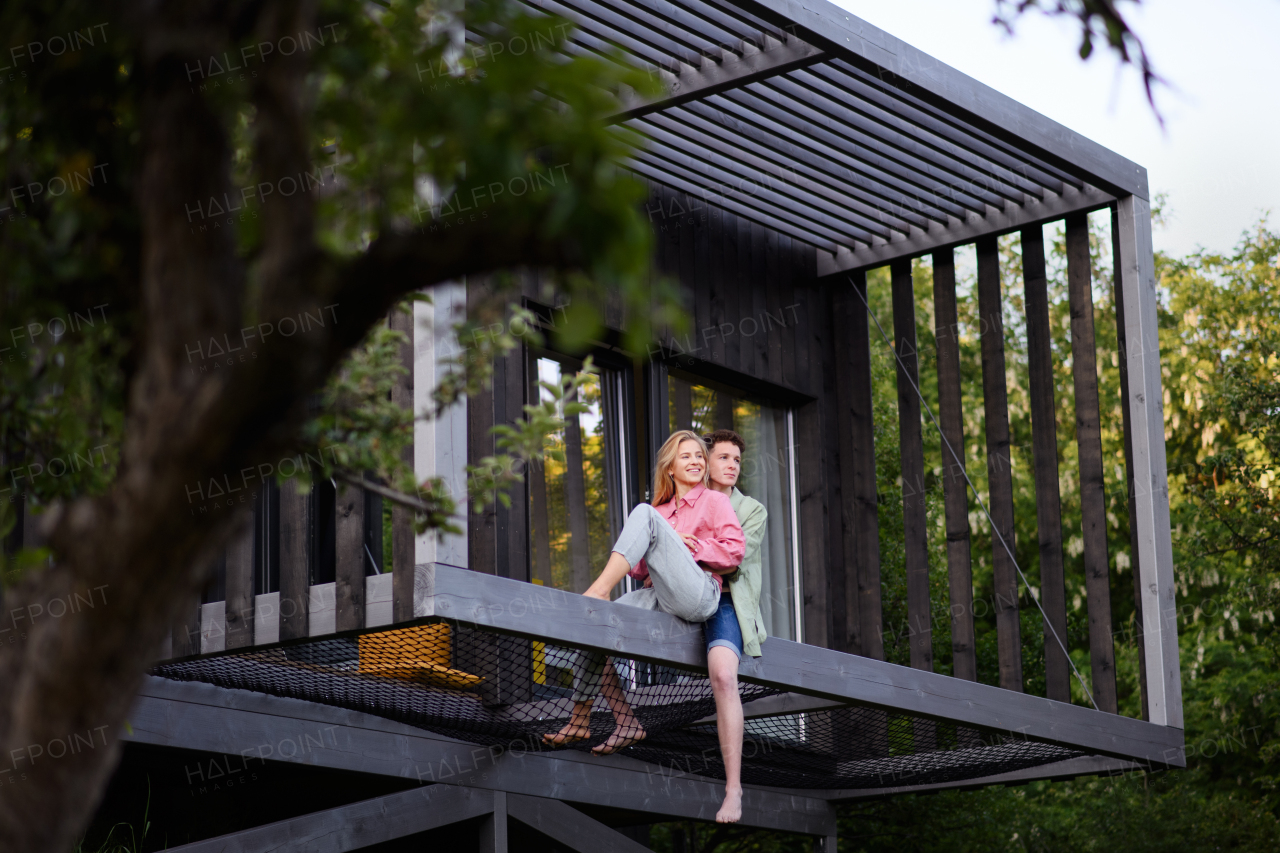 A young couple sitting and cuddling in hammock terrace in their new home in tiny house in woods, sustainable living concept.