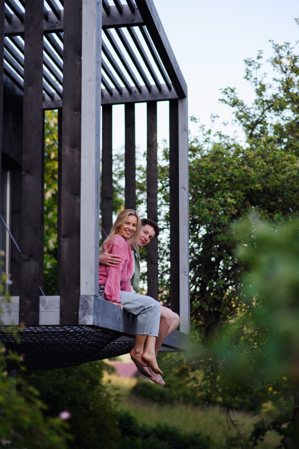 A young couple sitting and cuddling in hammock terrace in their new home in tiny house in woods, sustainable living concept.