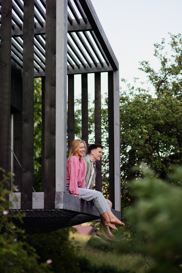 A young couple sitting and cuddling in hammock terrace in their new home in tiny house in woods, sustainable living concept.