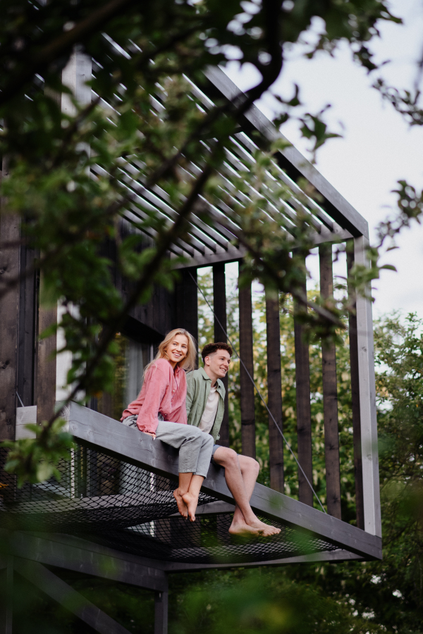 A young couple sitting and cuddling in hammock terrace in their new home in tiny house in woods, sustainable living concept.