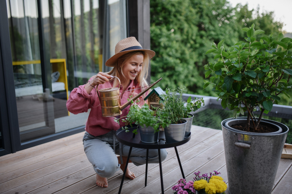 A young woman taking care of her herbs, watering them on terrace in tiny house, sustainable living.