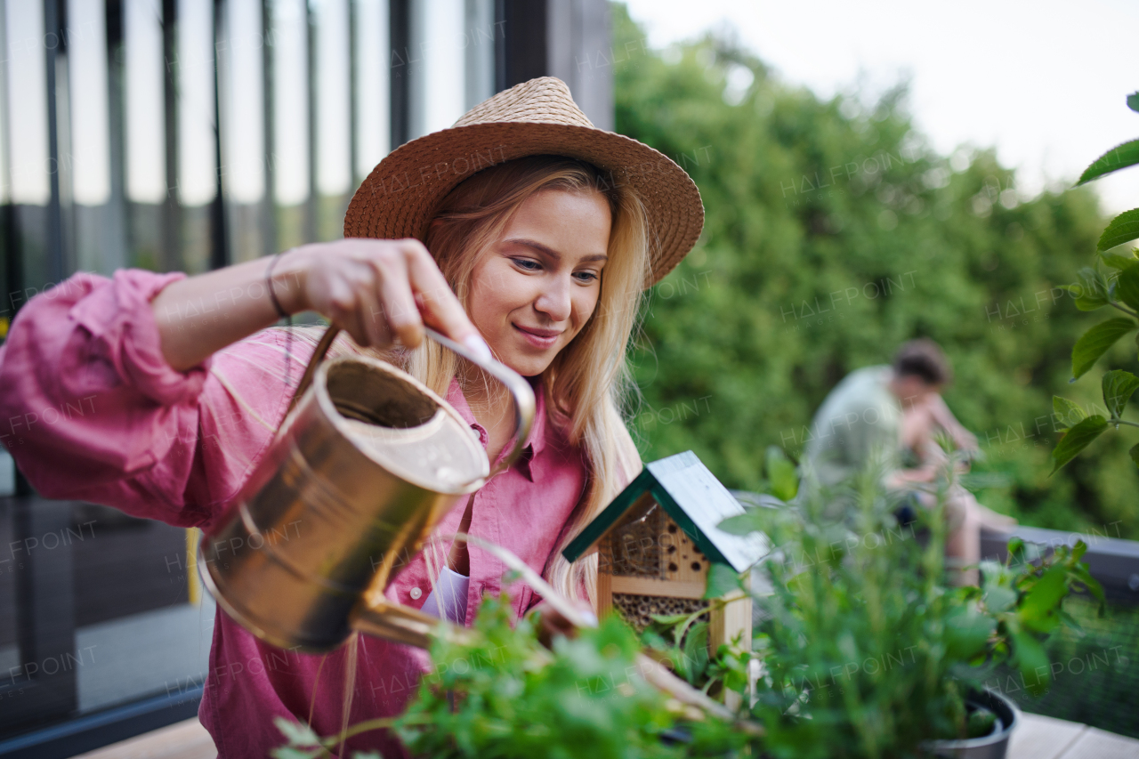 A young woman taking care of her herbs, watering them on terrace in tiny house, sustainable living.