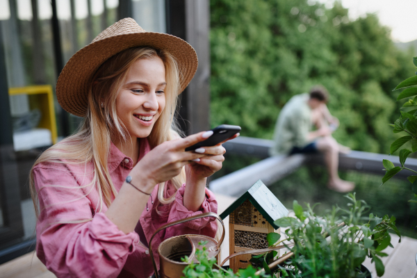 A young woman taking care of her herbs and taking picture of it on terrace in tiny house, sustainable living.