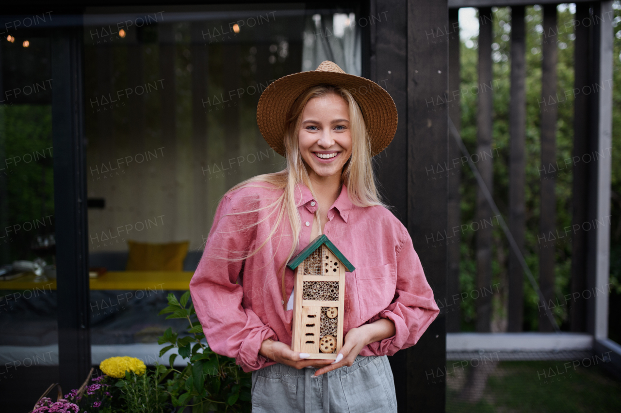 A young woman garedener holding bug and insect hotel on terrace in garden, sustainable lifestyle.