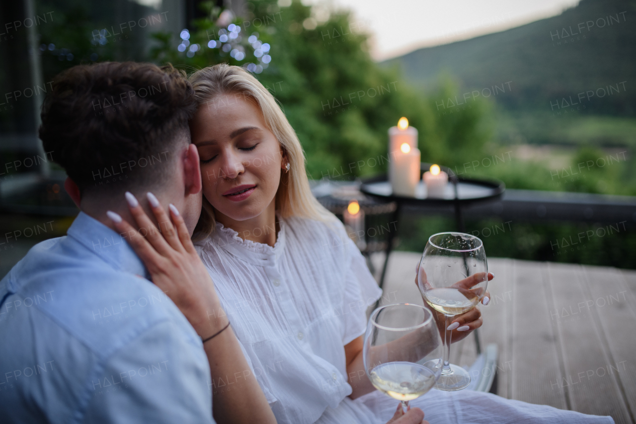 A young couple with wine resting outdoors on terrace in evening, weekend away in tiny house in countryside, sustainable living.