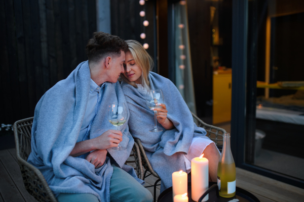 A young couple with wine resting outdoors on terrace in evening, weekend away in tiny house in countryside, sustainable living.