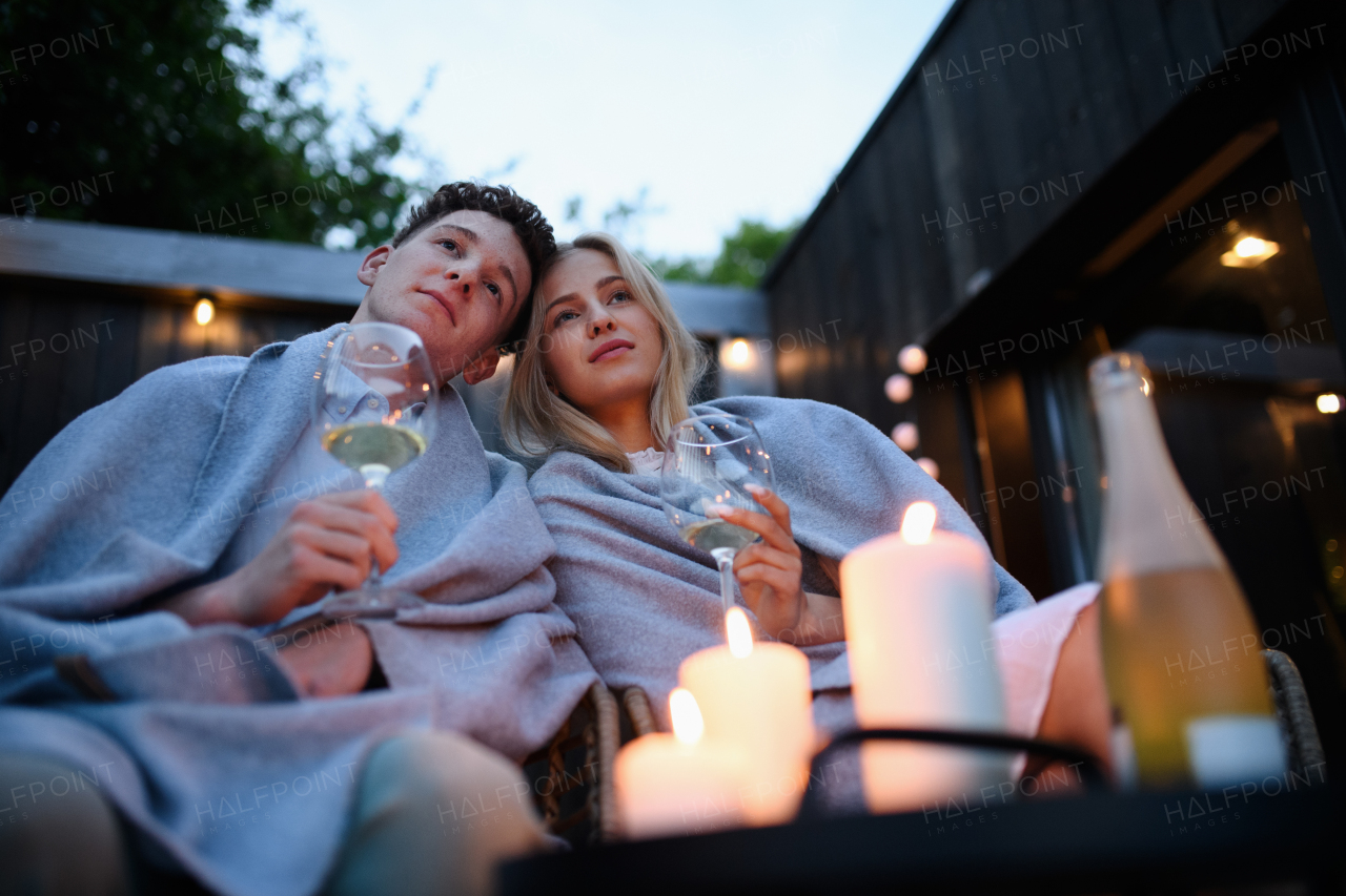 A young couple with wine resting outdoors on terrace in evening, weekend away in tiny house in countryside, sustainable living.