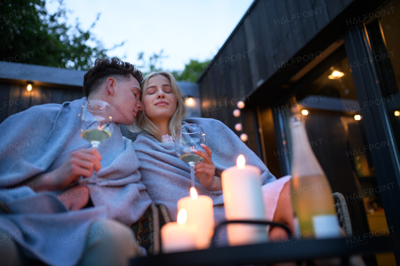 A young couple with wine resting outdoors on terrace in evening, weekend away in tiny house in countryside, sustainable living.